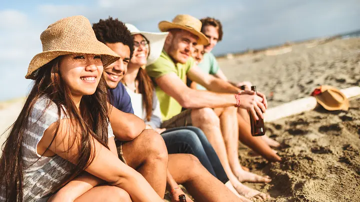 Gruppen-Reiseversicherungen - Eine Gruppe junger Menschen sitzen am Strand. Die Sonne schneit.