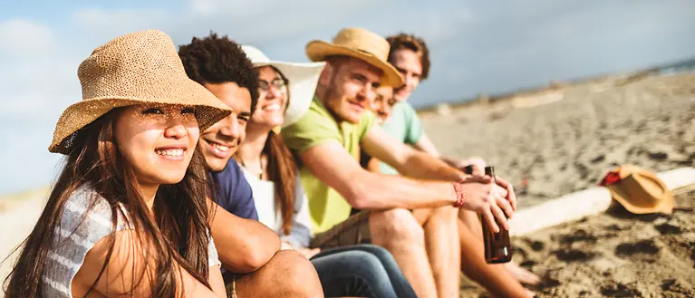 Gruppen-Reiseversicherungen - Eine Gruppe junger Menschen sitzen am Strand. Die Sonne schneit.