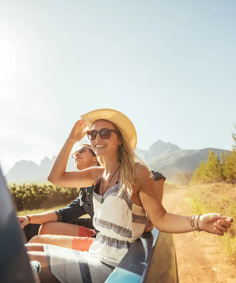 Eine Frau und ein Mann sitzen auf der Ladefläche eines Autos und genießen die Sonne. Im Hintergrund sind die Berge. 